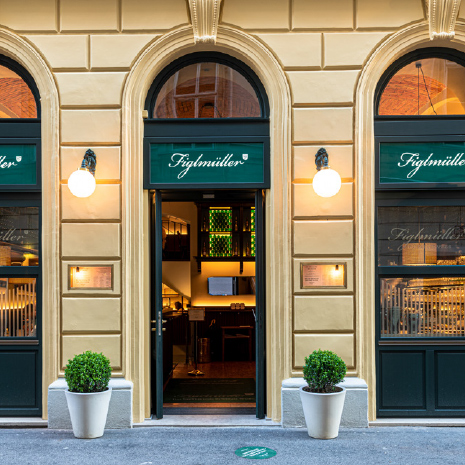 Straßenansicht des Eingangs vom Restaurant Figlmüller in der Bäckerstraße mit cremefarbener Fassade, Rundbögen und grüner Beschriftung.