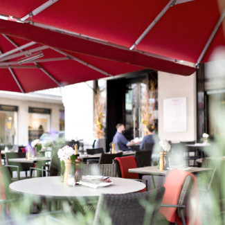 Straßenansicht des Gastgartens vom JOMA mit roten Sonnensegeln und Sitzplätzen.
